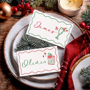 May include: Two place cards with names "James" and "Olivia" in red and green script, with festive illustrations. The cards are on a white plate with a red napkin and Christmas decorations, creating a holiday theme.