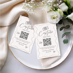 May include: Two cream-colored wedding cards with the text "Capture The Love" and a QR code. The cards also include the names "Olivia & Henry" and the date "04.12.26". The cards are on a white plate with floral decorations.