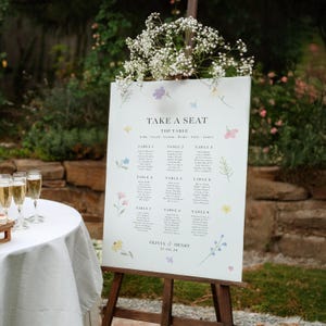 May include: A white wedding seating chart sign on a wooden easel, featuring the text TAKE A SEAT, TOP TABLE, and tables 1 through 9. The sign is decorated with small, colourful floral illustrations and the names Olivia & Henry with the date 21.08.26.