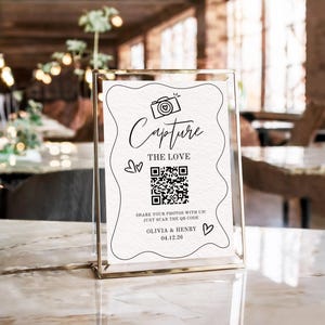 May include: A clear acrylic sign with a gold frame. The sign features the words "Capture The Love" with a camera illustration and a QR code. Below the QR code is the text "Share your photos with us! Just scan the QR code" and the names "Olivia & Henry" with the date 04.12.26.
