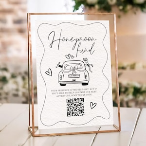 Puede incluir: Un letrero enmarcado con las palabras "Honeymoon Fund" en escritura elegante. El letrero presenta una ilustración en blanco y negro de un coche con una matrícula "Just Married", corazones y un código QR.