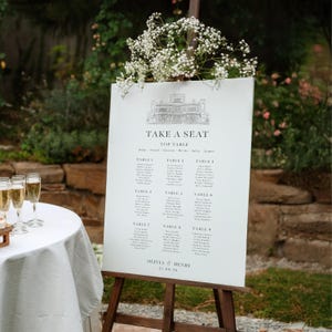 May include: A wedding seating plan displayed on a wooden easel, decorated with white flowers. The white sign features a building illustration, the words "TAKE A SEAT", and guest names arranged by table. The date is 21.09.26.