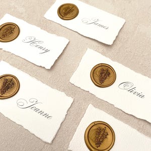 May include: Five white place cards with torn edges, each featuring a name in elegant script and a gold wax seal with a botanical design. Names include James, Henry, Joanne, and Olivia. The cards are arranged on a neutral surface.