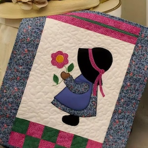 May include: A quilted wall hanging with a child figure holding a flower. The design has a white background, a blue floral border, and pink and green accents. The child wears a blue dress and a black bonnet. The quilt is likely handmade.