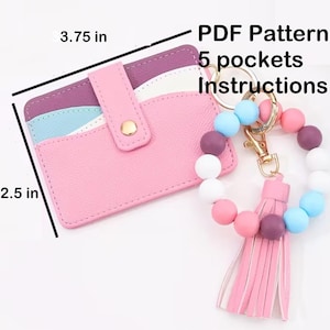 Può includere: Un porta carte rosa, largo 9,5 cm e alto 6,35 cm, con chiusura a scatto, accanto a un braccialetto di perline con nappa. Il porta carte ha più fessure colorate. Il braccialetto ha una chiusura dorata e una nappa rosa.