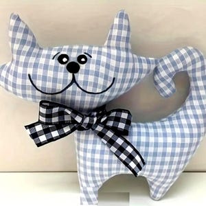 May include: A cat-shaped cushion made from blue and white gingham fabric. The cat features black embroidered eyes, nose, and a smiling mouth. A black and white checked bow is tied around its neck. The cat's tail is curved.