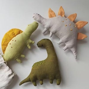 May include: Three soft dinosaur toys in shades of green, grey, and yellow. The toys have embroidered details and are arranged on a white surface. One dinosaur has a yellow sail, another has orange plates, and the third has a long neck.