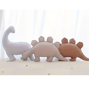 May include: Three plush dinosaur-shaped pillows in a row. The first is a light gray Brontosaurus, the second is a tan Stegosaurus, and the third is a brown Stegosaurus. All have stitched details and are on a textured surface.