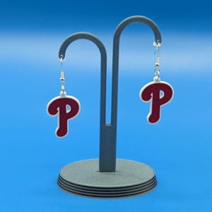 May include: Miniature earrings featuring a maroon "P" design with a white border, displayed on a gray stand against a blue backdrop. The earrings are approximately .82 inches wide and 1.18 inches long. The text "Mini Size" is displayed at the top.