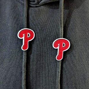 Peut inclure: Gros plan d'un sweat à capuche noir avec deux écussons brodés rouges et blancs en forme de lettre "P". Les écussons sont de chaque côté du cordon de serrage. Le sweat à capuche a des œillets métalliques.
