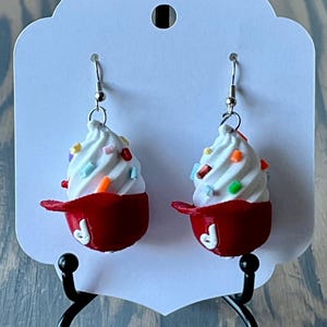 May include: A pair of dangle earrings featuring a red and white ice cream cone with sprinkles. The earrings are made of polymer clay and have silver hooks.