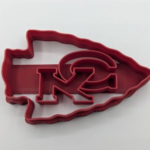 May include: Red plastic cookie cutter shaped like an arrowhead with the letters "KC" and a stylized "C" inside.