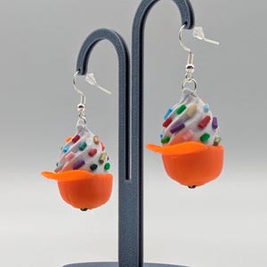 May include: A pair of novelty earrings shaped like miniature ice cream cones. Each earring features a white swirl of 'ice cream' with colorful sprinkles, set in an orange 'bowl' with a matching orange 'spoon'. The earrings hang from silver-colored hooks.
