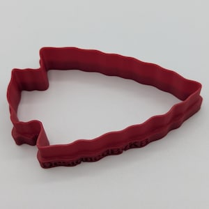 Peut inclure: Un emporte-pièce en plastique rouge en forme de pointe de flèche. Le découpoir a un bord ondulé et le texte "DesignedByKB" est imprimé le long du bord inférieur. Le découpoir est sur fond blanc.