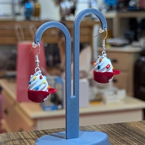 Op de afbeelding: Een paar novelty oorbellen in de vorm van miniatuur ijscoupes. Elke oorbel heeft een rode beker, wit ijs en kleurrijke hagelslag. De oorbellen hangen aan zilverkleurige haken en worden tentoongesteld op een blauwe oorbelstandaard.