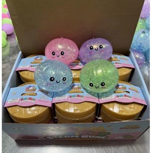 May include: A box of colorful squishy toy steam buns. The buns are in pink, purple, blue, and green, with glitter and smiling faces. Each bun sits atop a brown container with a blue and pink label that reads "Steam Buns".