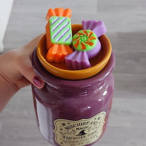 Könnte beinhalten: Ein violetter Keramik-Wachswärmer mit einem Etikett, auf dem MISCHIEF AND MAGIC TRICKSTER TONIC USE DURING A FULL MOON steht. Die obere Schale enthält zwei Wachsschmelzen in Form von Bonbons, eines in Orange und Grün, das andere in Violett und Grün.