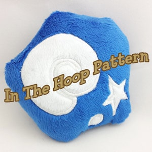 Puede incluir: Juguete de peluche azul con forma de concha marina con un remolino blanco bordado y una estrella blanca. El texto "In The Hoop Pattern" está bordado en el juguete.