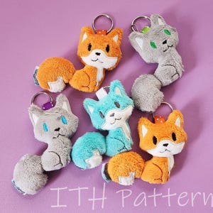 May include: Five plush animal keychains, including a grey cat, a blue fox, an orange fox, and two orange foxes. The keychains are all made of soft, fuzzy fabric and have embroidered details. The keychains are arranged in a circle on a purple background.