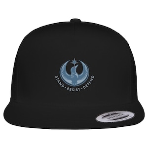 May include: Black trucker cap with a blue and white bird and star emblem, and the words "STAND. RESIST. DEFEND." The cap has a curved peak and a snapback fastening.