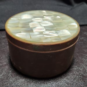 Può includere: Una piccola scatola rotonda marrone con un coperchio intarsiato con piastrelle iridescenti di madreperla. La scatola ha un design semplice ed elegante, adatto per riporre piccoli oggetti. La superficie del coperchio è un mosaico di pezzi rettangolari.