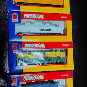 Puede incluir: Cuatro cajas de modelos de trenes de carga a escala HO. Las cajas son rojas, amarillas y azules con la inscripción "FREIGHT CAR". Las cajas contienen modelos de vagones de tren con los nombres "CHICA GO", "Potlatch", "CNW" y "L & N".