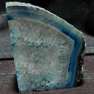 May include: A polished, cut agate stone with bands of blue, gray, and white. The natural stone has a crystalline interior and a dark blue outer edge. The surface is textured with natural patterns and variations.