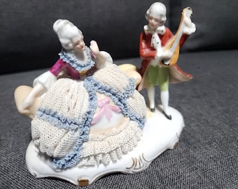 May include: A porcelain figurine set featuring a seated figure in a magenta bodice and a cream-colored skirt with blue lace trim, accompanied by a standing figure in a red coat and green breeches playing a lute, all on a white base.