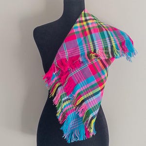 May include: A colorful plaid scarf with a fringed edge. The scarf features a vibrant mix of pink, blue, yellow, and green in a classic plaid pattern. The scarf is draped over a black mannequin.