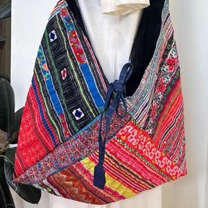 May include: A colorful patchwork shoulder bag with a black strap. The bag features vibrant patterns in red, blue, green, and yellow. The strap is a dark blue fabric with a patterned design. The bag is tied with a blue cord.