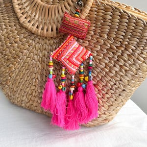 May include: Woven straw tote bag with a rounded handle. Attached are two decorative charms: a small embroidered rectangle and a square with pink tassels and colorful beads. The bag and accessories are in shades of tan, pink, red, and yellow.