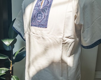 May include: Off-white short-sleeved shirt with a blue embroidered design on the chest and collar. The shirt has a relaxed fit and is displayed on a mannequin. The design features geometric patterns.