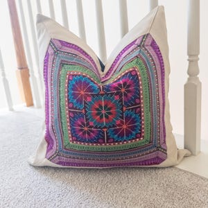 May include: A decorative square pillow with a colorful, geometric design. The pillow features a central floral pattern in shades of blue, pink, and green, framed by a border of purple and green stripes. The pillow is set against a neutral background.