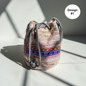 May include: A beige drawstring bag with a colorful embroidered pattern. The bag features a gray rope drawstring and is decorated with pink, blue, and green floral and geometric designs. The text "Design #1" is in a white circle.