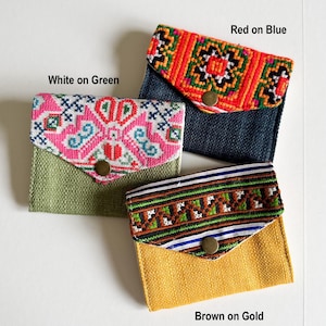 May include: Three small, colorful fabric wallets with patterned flaps. One is white on green, another red on blue, and the third brown on gold. Each wallet has a button closure. The text "White on Green", "Red on Blue", and "Brown on Gold" are visible.