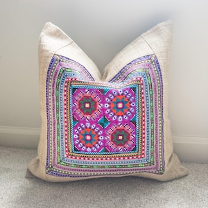 May include: Beige square pillow with a vibrant, embroidered design. The central pattern features four magenta floral shapes with teal and orange accents. The border showcases a colorful geometric pattern in shades of purple, green, and blue. The pillow is resting on a gray carpet.