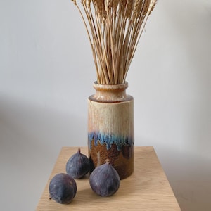 May include: A decorative ceramic vase with a beige, brown, and blue glaze, filled with dried wheat stalks. Three ripe figs are placed in front of the vase on a wooden surface. The vase is cylindrical with a flared rim.