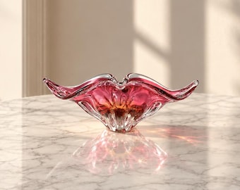 Può includere: Una ciotola decorativa in vetro caratterizzata da un vivace gradiente di tonalità magenta intenso e ambra. La ciotola presenta un bordo ondulato distintivo con lati allungati e appuntiti e una base in vetro trasparente con finitura liscia.