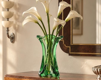 Può includere: Un vaso in vetro verde vibrante con un bordo svasato e ondulato e una base spessa e modellata. Il vaso contiene un bouquet di sei fiori di calla bianchi con lunghi steli verdi, creando un contrasto sorprendente tra il vetro verde e i fiori bianchi.