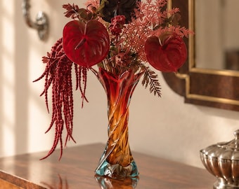 May include: A tall, twisted glass vase with a gradient of red, orange, and blue hues, holding a floral arrangement of deep red anthurium flowers, dark foliage, and long, drooping red amaranth stems, creating a vibrant and textured display.