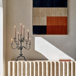 Könnte beinhalten: Ein schwarzer Metallkandelaber mit sieben brennenden Stabkerzen, ein rechteckiges Wandbild mit horizontalen Blöcken in Marineblau, Beige und gebranntem Orange sowie ein beiger, geriffelter Konsolentisch mit einem kleinen rechteckigen Objekt.