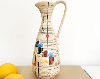 May include: A beige ceramic pitcher with a handle, decorated with black vertical lines and colorful geometric shapes. The pitcher has a slender neck and a wide base. Two yellow lemons are in the foreground.