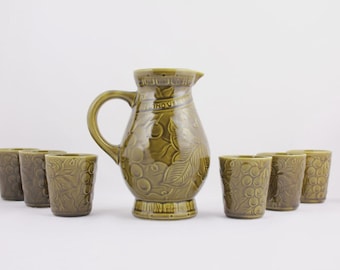 May include: A green ceramic pitcher and six matching glasses. The pitcher has a handle and a spout, and the glasses are small and cylindrical. All pieces feature a grape and leaf design.