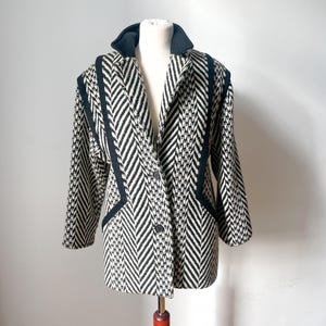 May include: A black and white patterned coat with a black collar and trim. The coat is displayed on a mannequin. The pattern is a mix of herringbone and other geometric designs. The coat has two buttons.