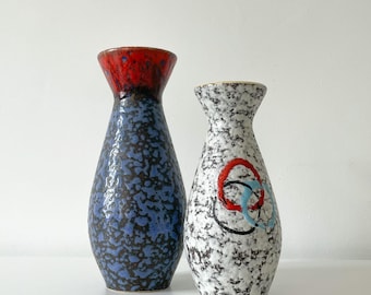 May include: Two ceramic vases. The vase on the left is blue with a red top. The vase on the right is white with black speckles and a red, black, and blue design. Both vases have a textured surface.