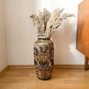 Op de afbeelding: Een hoge, cilindrische keramische vaas met een textuurrijke, olijfgroene glazuurlaag, voorzien van reliëfbloem- en hartmotieven in oranje, blauw en donkerbruin. De vaas is gevuld met verschillende stengels gedroogd, pluizig, beige pampasgras.