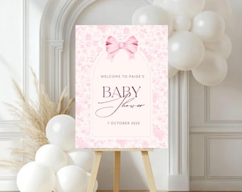 May include: A pink baby shower sign on a wooden easel. The sign features a floral pattern, a pink bow, and the words "Welcome to Paige's Baby Shower 7 October 2025."