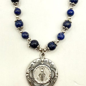 Puede incluir: Un collar con cuentas de lapislázuli azul intenso separadas por pequeños espaciadores de metal plateado y casquillos ornamentados. Un colgante circular plateado muestra una figura con el texto O MARY CONCEIVED WITHOUT SIN.