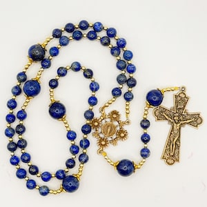 Può includere: Un rosario con perline di lapislazzuli blu scuro e dettagli dorati. Il design include un crocifisso e un elemento centrale decorativo con un emblema religioso. Le perline sono collegate da filo dorato.