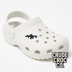 May include: White clog shoe with a black gun-shaped charm. The shoe has multiple holes and a pivoting heel strap. The text "CRUDE CROC CO." is printed in black and white on the lower right side.